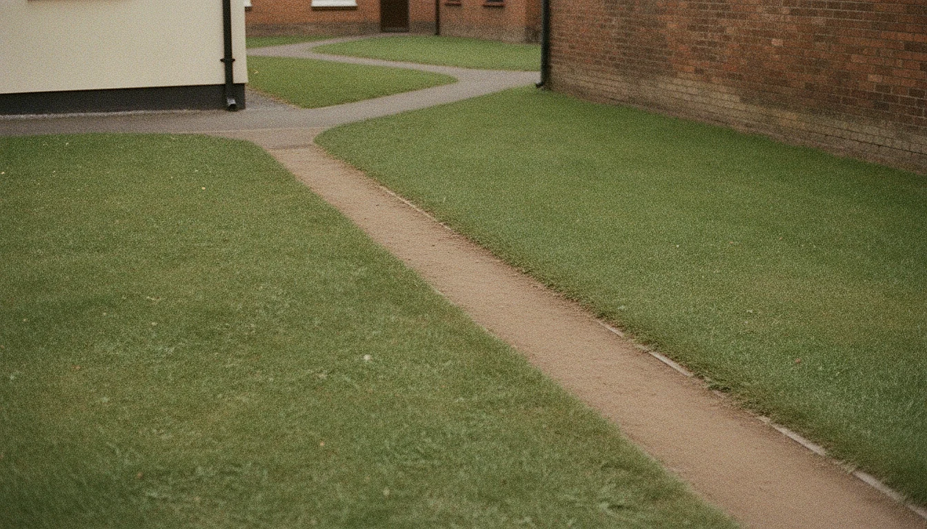 A narrow dirt path worn diagonally across a grass courtyard between two ordinary buildings.